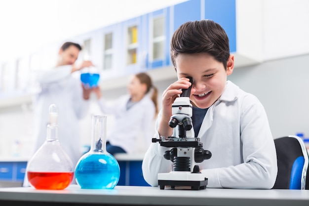 Muestra interesante. Niño feliz feliz creativo usando microscopio mientras sonríe y usa bata de laboratorio