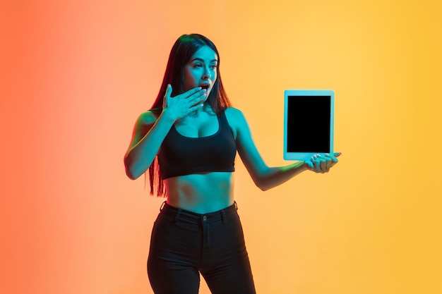 Muestra la pantalla de la tableta. Retrato de joven mujer caucásica sobre fondo de estudio degradado amarillo-naranja en luz de neón. Concepto de juventud, emociones humanas, expresión facial, ventas, publicidad. Modelo de ajuste deportivo.