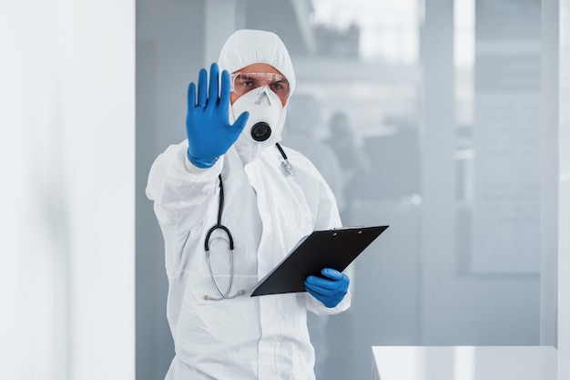 Muestra parada gesto con la mano. Doctor científico masculino en bata de laboratorio, gafas defensivas y máscara con bloc de notas en las manos