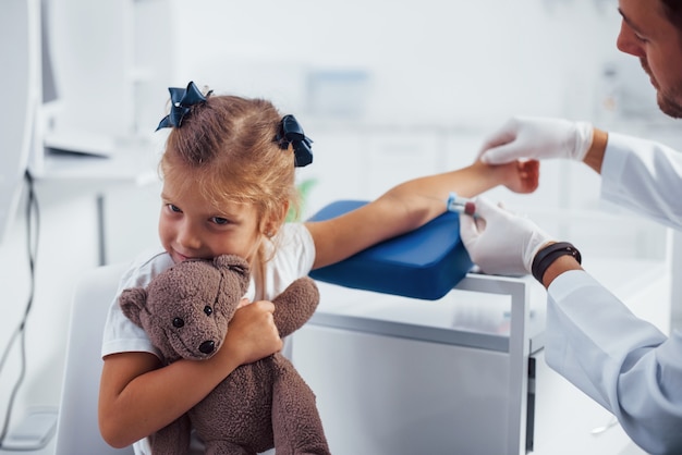 Muestra de sangre. La niña con su osito de peluche está en la clínica con el médico.