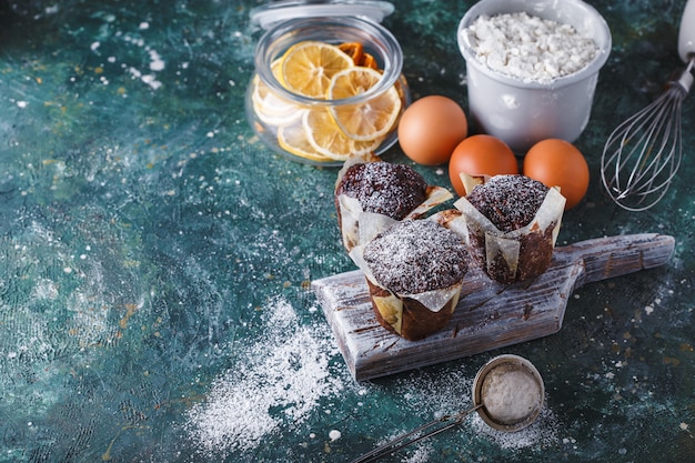 Muffin de cenoura e chocolate polvilhado com açúcar de confeiteiro, uma xícara de chá, ingredientes de panificação. Farinha, ovos, limão cítrico em uma mesa escura