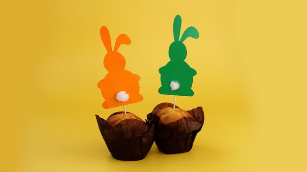 Muffin decorado con un conejo de papel en un palillo. Decoración de Pascua para cupcakes. Dulces y repostería festiva. Fondo amarillo