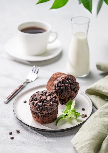 Muffins de chocolate en un plato blanco con menta en un fondo claro con una taza de leche de café y rama