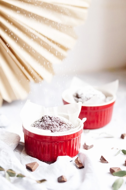 Muffins de chocolate en tazas rojas. Pequeña cazuela de cerámica vidriada con tortas marrones sobre fondo gris y blanco.