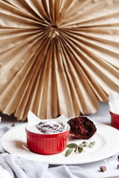 Muffins de chocolate en tazas rojas. Pequeña cazuela de cerámica vidriada con tortas marrones sobre fondo gris y blanco.