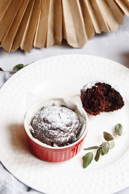 Muffins de chocolate en tazas rojas. Pequeña cazuela de cerámica vidriada con tortas marrones sobre fondo gris y blanco.