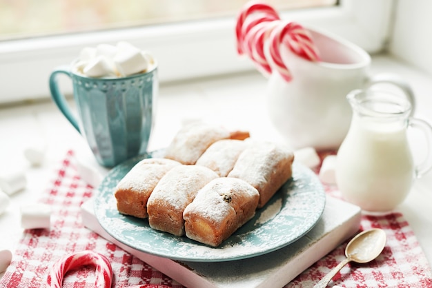 Muffins navideños, leche, cacao, malvaviscos, paletas de caramelo en un plato blanco junto a la ventana