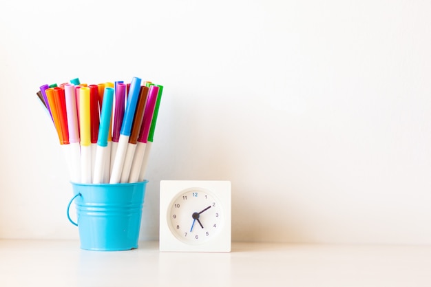 Foto muitas canetas de feltro multicoloridas em suporte de cesta azul com pequeno relógio branco na mesa isolado fundo branco, espaço de cópia