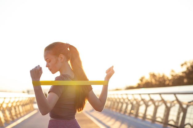 Muito apto mulher com cabelo ruivo, vestida com roupas esportivas, fazendo exercícios com elástico na ponte pela manhã. Espaço para texto