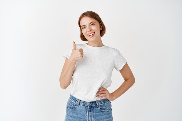 Muito bem. modelo feminino sorridente em t-shirt mostrando o polegar para cima em aprovação. mulher jovem com aparência natural, elogio e gosto do produto, satisfeita em pé na parede branca