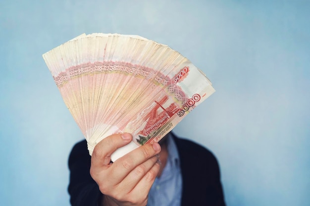 Muito dinheiro na mão de um jovem empresário em um fundo azul Uma pilha de notas de rublos russos com um valor nominal de 5000 rublos empresário de sucesso