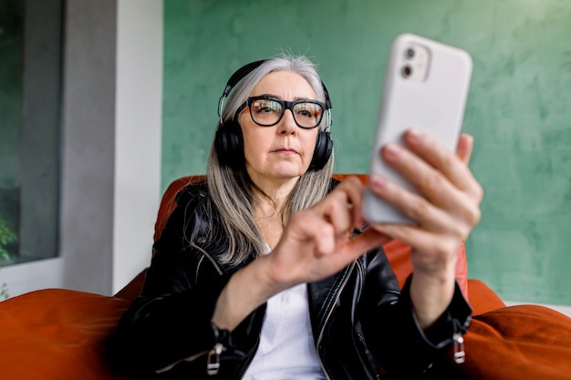 Muito elegante senhora sênior de óculos com cabelos grisalhos lisos que sentado na cadeira vermelha e ouvindo música bonita em fones de ouvido de sua lista de reprodução de smartphones