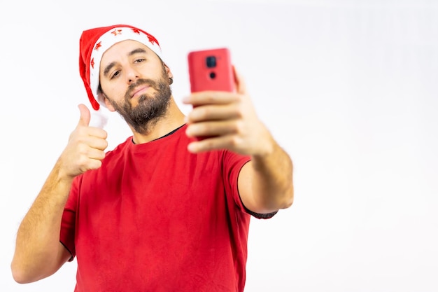 Muito feliz, jovem homem branco com o chapéu vermelho de Natal tirando uma selfie em um fundo branco, vestido com uma camiseta vermelha, copiar e colar