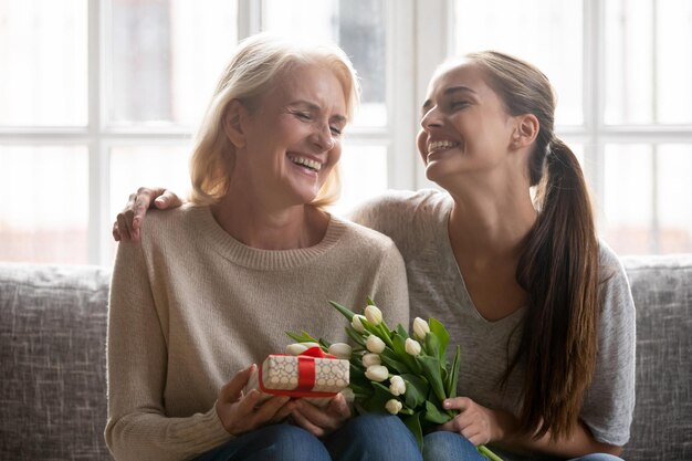 Muito feliz, linda mãe de meia-idade senta-se com a filha crescida segurando presentes e flores família rindo comemorando aniversário de evento de vida ou feriado de primavera 8 de março Dia Internacional da Mulher se sentindo feliz