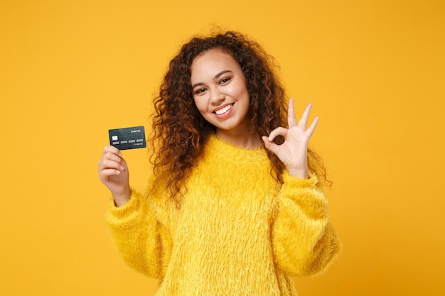 Muito jovem afro-americana em suéter de pele posando isolado no retrato de estúdio de fundo laranja amarelo. conceito de estilo de vida de pessoas. mock up espaço de cópia. segure o cartão de crédito, mostrando o gesto de ok.