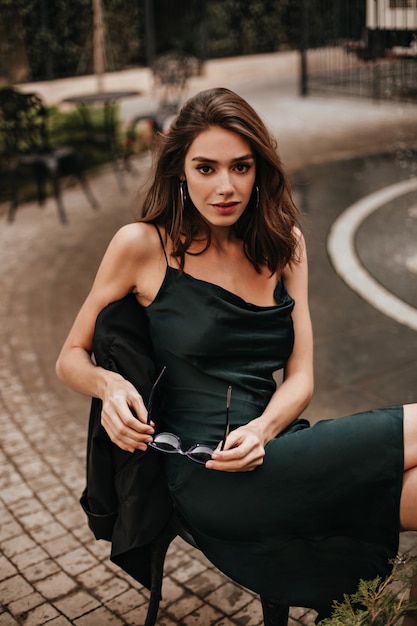 Muito jovem com cabelo ondulado morena, maquiagem brilhante e vestido midi verde, posando na cadeira no terraço do café da cidade e olhando para a frente