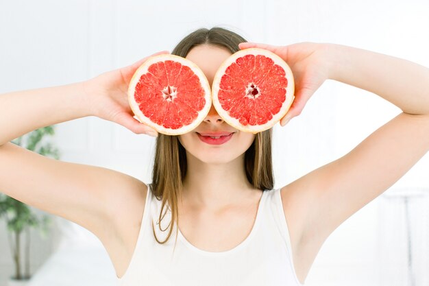 Muito jovem com fatias de laranja na frente de seus olhos. Linda mulher segurando a fatia de toranja, escondendo os olhos atrás isolado no fundo branco