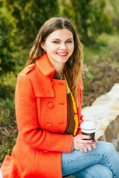 Muito jovem de casaco vermelho com uma xícara de café feliz andando no parque