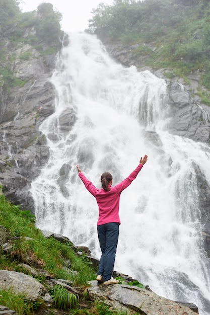 Muito jovem de pé com as mãos levantadas na grande cachoeira poderosa. mulher bonita, curtindo a natureza em clima quente de outono