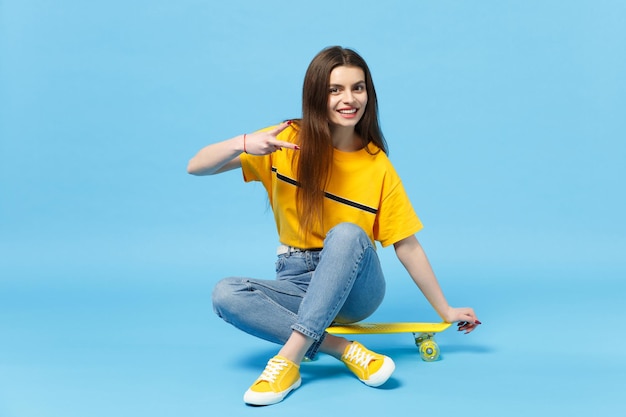 Muito jovem em roupas casuais vívidas, olhando para a câmera, mostrando o sinal de vitória sentado com skate amarelo isolado no fundo da parede azul no estúdio. Conceito de estilo de vida de pessoas. Mock up espaço de cópia.