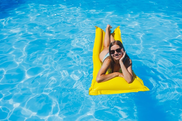 Muito jovem em traje de banho e óculos de sol deitado em colchões infláveis amarelos na piscina Desfrute de banhos de sol e relaxamento