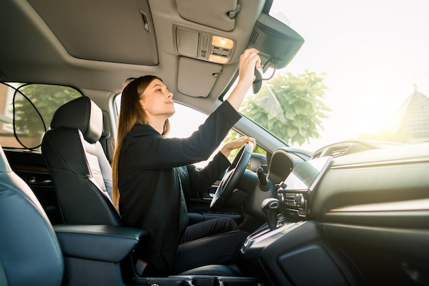 Muito jovem empresária caucasiana com roupa formal, sentado no banco do motorista no carro dela e verificando o espelho retrovisor central