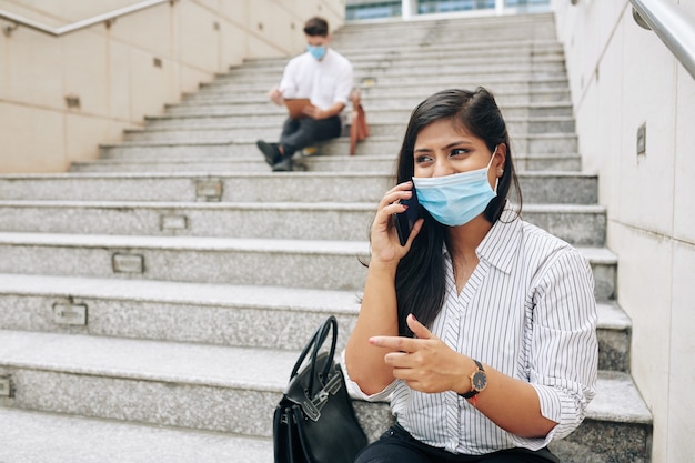 Muito jovem empresária emocional em máscara médica sentada nos degraus e falando ao telefone com um colega