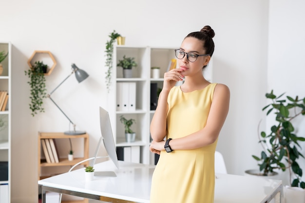 Muito jovem empresária pensativa em um elegante vestido amarelo e óculos no local de trabalho no escritório