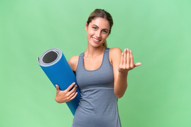 Muito jovem esporte uruguaio indo para aulas de ioga enquanto segura um tapete sobre fundo isolado convidando para vir com a mão feliz que você veio