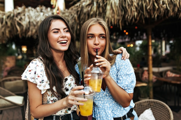 Foto muito jovem morena com blusa floral branca e mulher loira atraente bronzeada com top azul beber uma limonada saborosa lá fora