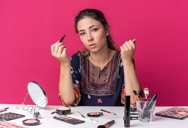 Foto muito jovem morena sentada à mesa com ferramentas de maquiagem segurando um delineador