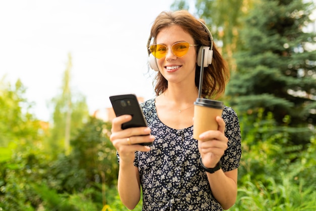 Muito jovem mulher caucasiana dar um passeio no parque com café e fones de ouvido