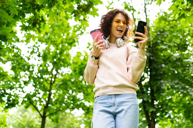 Muito jovem mulher caucasiana dar um passeio no parque com uma xícara de café e se comunicar com os amigos com seu telefone