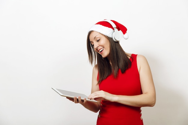 Muito jovem mulher feliz em um vestido vermelho e chapéu de natal ouvindo música em fones de ouvido, segurando o tablet em fundo branco. gadget de menina santa isolado. feriado de ano novo 2018. copie o anúncio de espaço.