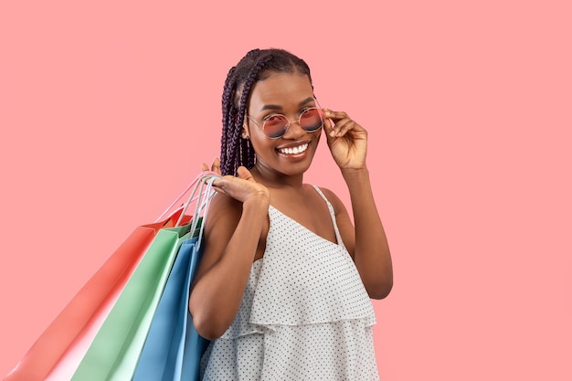 Muito jovem negra de vestido e óculos de sol segurando sacolas de compras sorrindo para a câmera em rosa