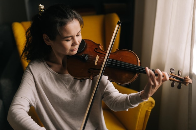 Muito jovem tocando violino closeup sentado na cadeira macia na sala com um interior moderno Garota está praticando tocando instrumento musical em casa