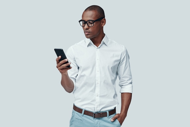 Muito ocupado. africano jovem e bonito usando um telefone inteligente em pé contra um fundo cinza