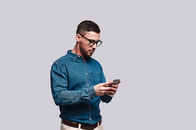 Muito ocupado. Jovem bonito em traje esporte fino usando seu telefone em pé contra um fundo cinza