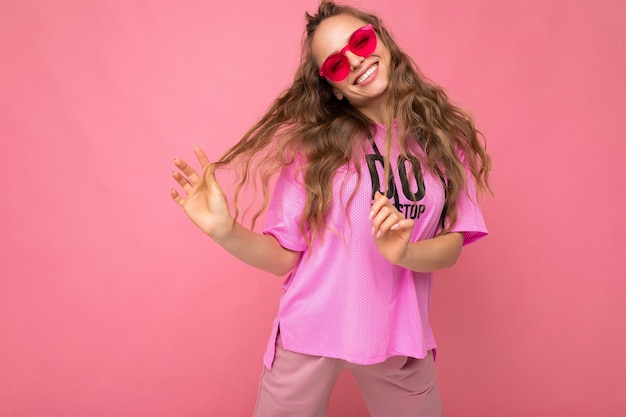 Muito positivo, sorridente jovem loira encaracolada isolada sobre a parede de fundo rosa, vestindo uma camiseta rosa casual e elegantes óculos de sol coloridos, olhando para a câmera