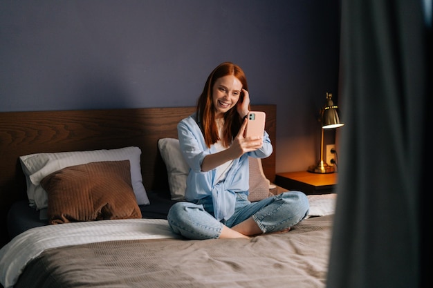 Muito ruiva jovem posando para selfie móvel na cama enquanto está sentado na cama no quarto aconchegante. Mulher caucasiana com videochamada no aplicativo de celular em casa.