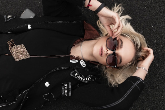 Muito sexy linda jovem de blazer preto elegante em óculos de sol da moda está descansando no asfalto na rua num dia de verão. Linda garota moderna relaxando na cidade