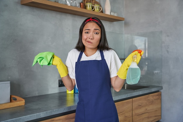 Muito trabalho, jovem faxineira olhando confusa para a câmera enquanto segura o spray de detergente