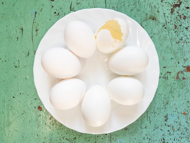 Muitos ovos brancos inteiros e um ovo quebrado estão em um círculo, em um prato branco sobre um fundo verde.