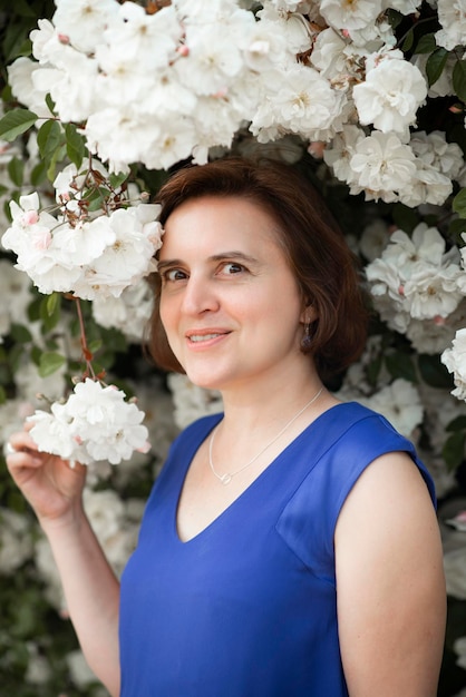 Mujer de 48 años con un vestido azul cerca de un floreciente rosal blanco