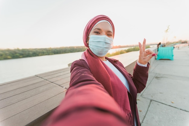 Mujer en abaya y máscara médica protectora grabando video en la cámara selfie mientras pasa tiempo en la calle