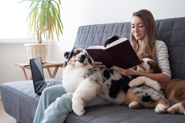 Mujer abrazando besando a tres Pequeño lindo pastor australiano rojo azul merle cachorro de perro en el sofá. Rojo Tres colores. leer cuaderno. Trabajar portátil en línea. Videollamada. Las compras en línea. Oficina en casa.