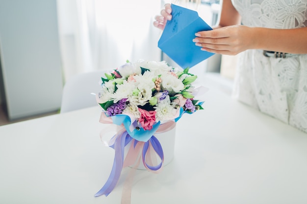 La mujer abre el sobre con la tarjeta dejada en ramo de flores en caja de regalo en cocina. Sorpresa. Presente para vacaciones