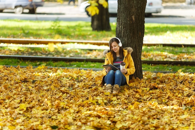 Una mujer con abrigo amarillo y jeans sentada con una taza de café o té y escuchando música debajo de un árbol con una tableta en sus manos y auriculares en el parque de la ciudad de otoño en un día caluroso. Otoño de hojas doradas.