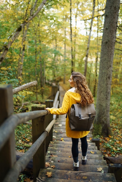 Mujer con un abrigo amarillo y mochila camina por una escalera de madera disfrutando del paisaje otoñal en el bosque