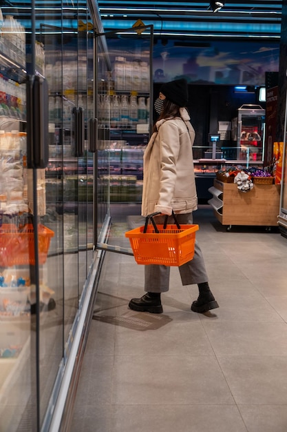 Mujer en abrigo y mascarilla médica en la tienda de abarrotes abriendo la puerta del frigorífico